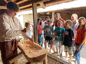 Kinder schauen einem Handwerker beim Aushöhlen eines Holztrogs zu